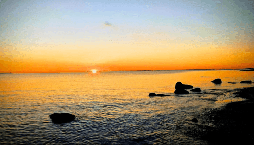 Sunset over calm waters, with rocks along the shore and a warm orange sky reflecting on the surface.