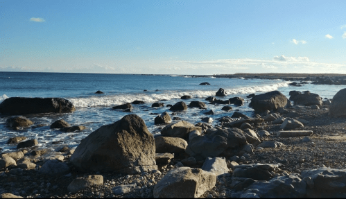 A rocky shoreline with gentle waves under a clear blue sky and distant clouds.