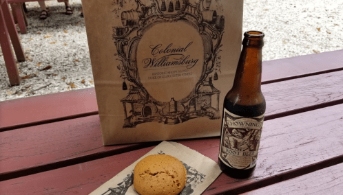 A brown paper bag from Colonial Williamsburg, a bottle of root beer, and a cookie on a wooden table.