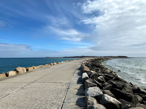 A rocky jetty extends into the ocean under a partly cloudy sky, with calm waters on either side.