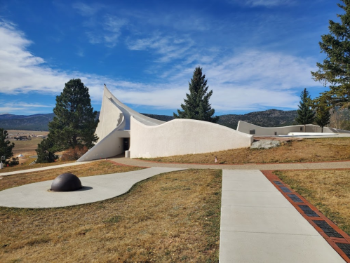 A modern, curved white building set against a blue sky, surrounded by trees and grassy landscape.