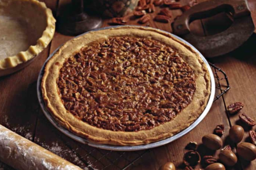 A freshly baked pecan pie on a wooden table, surrounded by pecans and a pie crust.