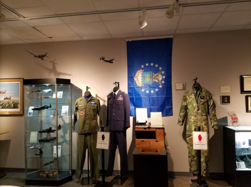 Display of military uniforms and artifacts, featuring a blue flag and model aircraft in a museum setting.