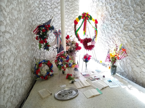 A memorial display featuring floral arrangements, flags, and open books on a textured surface.