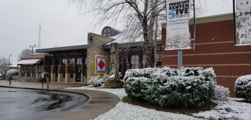 Snow covers the ground outside the Museum Center, showcasing a brick building with a colorful sign and greenery.