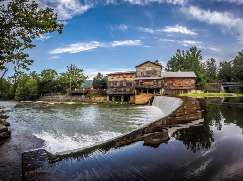 A historic wooden mill beside a calm river, with a small waterfall and lush greenery under a blue sky.