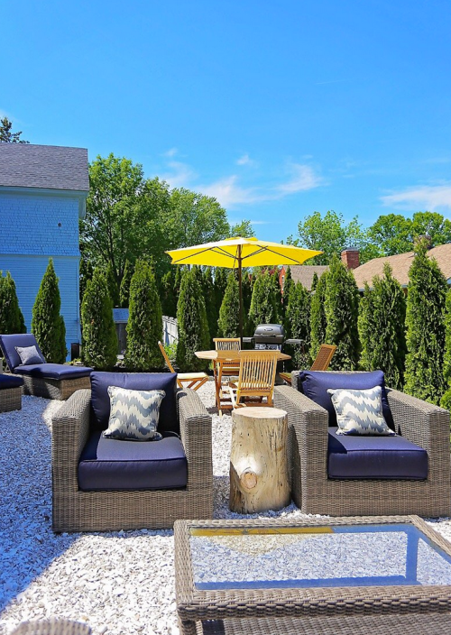 A cozy outdoor patio with wicker chairs, a yellow umbrella, and a wooden table surrounded by greenery and gravel.