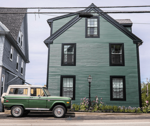 A green house with black trim and windows, next to a vintage green and yellow SUV parked on the street.