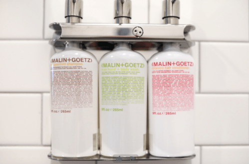 Three MALIN+GOETZ bottles in a wall-mounted holder: shampoo, body wash, and hand wash, against a tiled background.