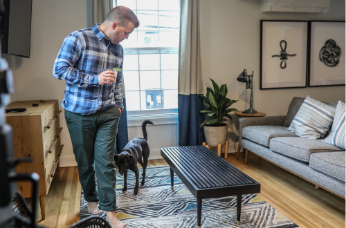 A man in a plaid shirt walks with a drink, accompanied by a black dog, in a cozy living room with modern decor.