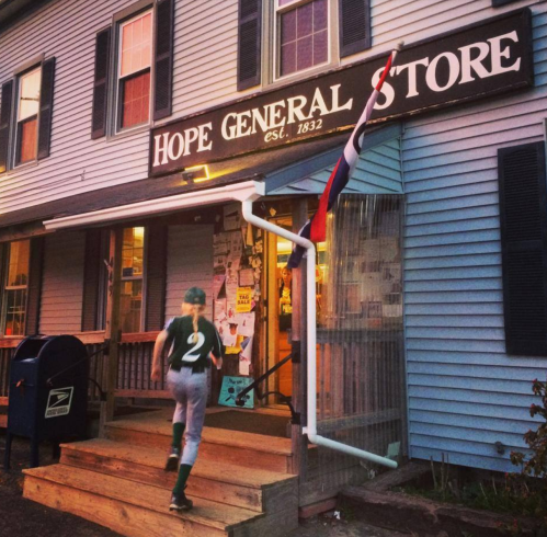 A child in a green sports uniform runs up the steps of Hope General Store, with a flag and mailbox nearby.