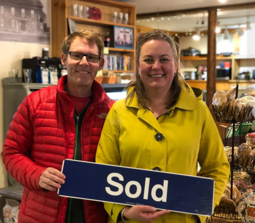 A man in a red jacket and a woman in a yellow coat hold a "Sold" sign in a cozy café setting.