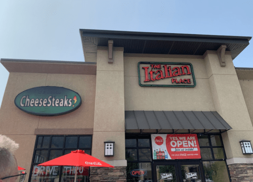 Exterior of a restaurant featuring signs for "CheeseSteaks" and "The Italian Place," with outdoor seating and umbrellas.