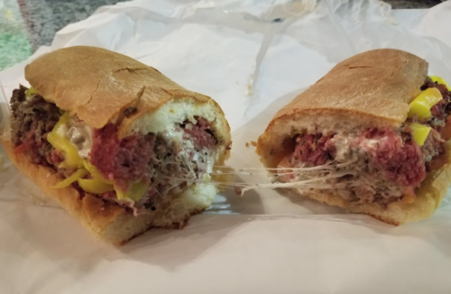 A close-up of a sandwich cut in half, filled with meat, melted cheese, and yellow peppers, resting on white paper.