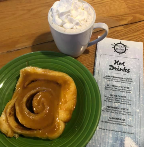 A cinnamon roll on a green plate next to a whipped cream-topped hot drink and a menu for hot beverages.