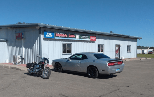A silver car parked next to a motorcycle in front of a blue building with signs for Bud Light and Captain's Cabin.