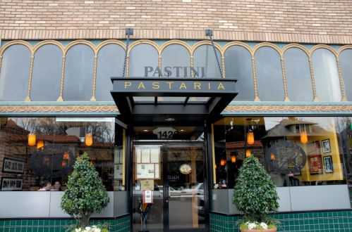 Exterior of Pastini Pastaria restaurant, featuring large windows, decorative lights, and potted plants at the entrance.