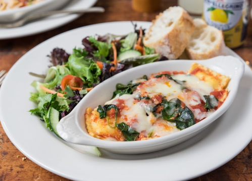 A dish of baked lasagna with spinach and cheese, served with a side salad and bread on a wooden table.