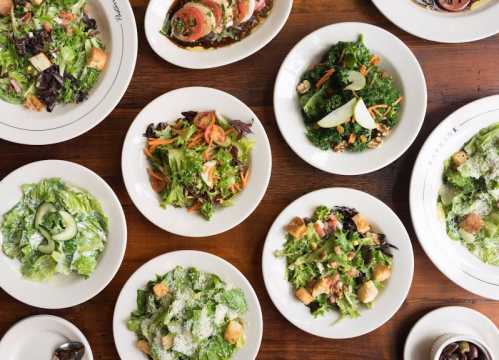 A variety of fresh salads served in white bowls on a wooden table, featuring greens, toppings, and dressings.