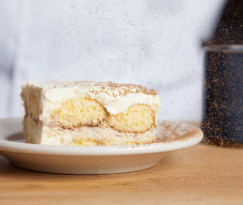 A slice of creamy dessert with layers of cake, topped with a dusting of cocoa powder on a white plate.