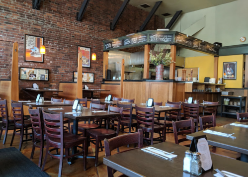 Cozy restaurant interior with wooden tables, chairs, and exposed brick walls, featuring a counter and decorative plants.
