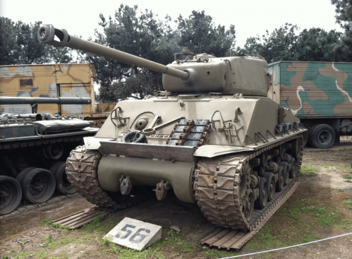 A military tank displayed outdoors, featuring a long barrel and tracks, set against a backdrop of trees and vehicles.