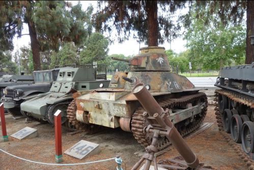 A vintage tank displayed outdoors among other military vehicles, surrounded by trees and gravel.