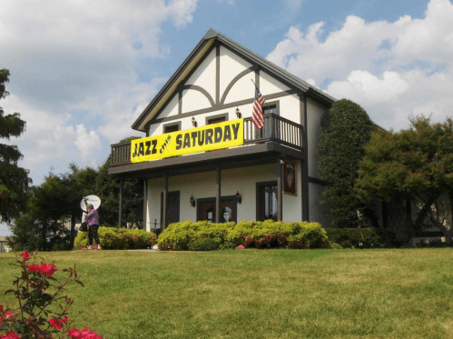 A two-story house with a large "JAZZ SATURDAY" banner, surrounded by greenery and flowers, under a partly cloudy sky.