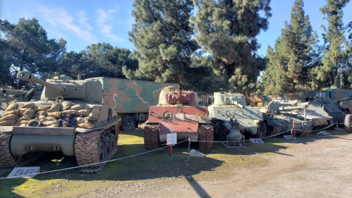 A row of military tanks displayed outdoors, surrounded by trees and a camouflage truck in the background.