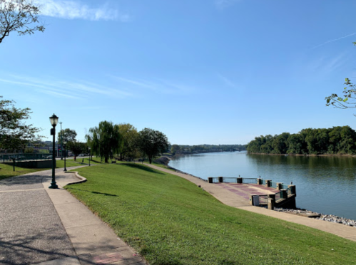 A serene riverside park with a walkway, green grass, trees, and calm water under a clear blue sky.