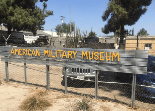 Sign for the American Military Museum, displayed on a fence with trees and vehicles in the background.