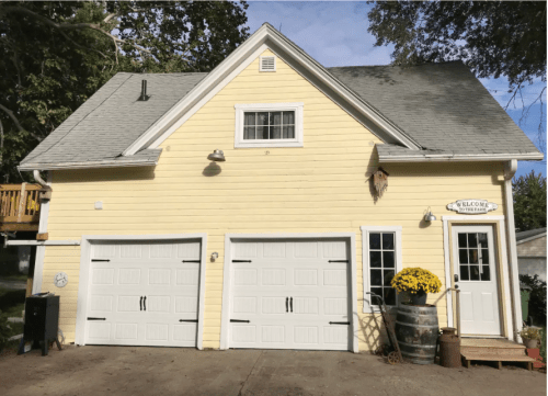 A yellow house with a steep roof, two garage doors, and a flower pot on the porch, set against a clear blue sky.