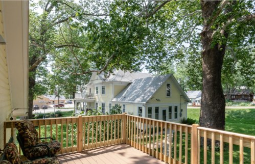View from a balcony overlooking a green yard and two houses, surrounded by large trees on a sunny day.