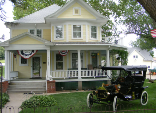 A yellow Victorian-style house with a vintage car parked in front, decorated with bunting and surrounded by greenery.