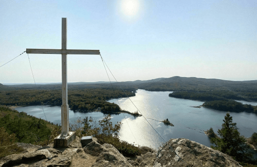 A wooden cross on a rocky outcrop overlooking a serene lake and forested hills under a clear blue sky.