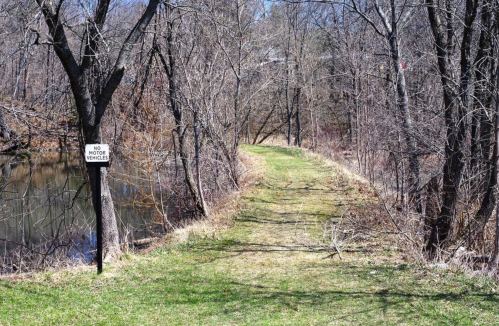 A narrow, grassy path along a river, bordered by bare trees and a sign that reads "No Motor Vehicles."