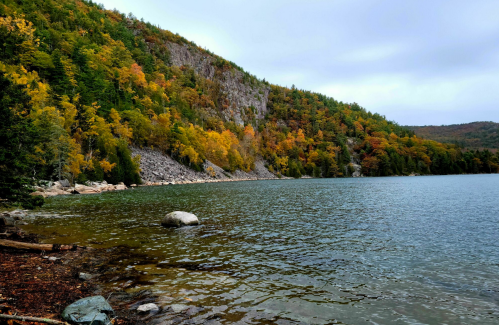 A serene lake surrounded by autumn-colored trees and rocky shores under a cloudy sky.