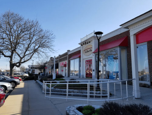 A shopping plaza with a red and white storefront, trees, and parked cars under a clear blue sky.