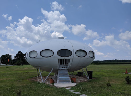 A futuristic, oval-shaped house with large windows, set in a grassy field under a blue sky with fluffy clouds.