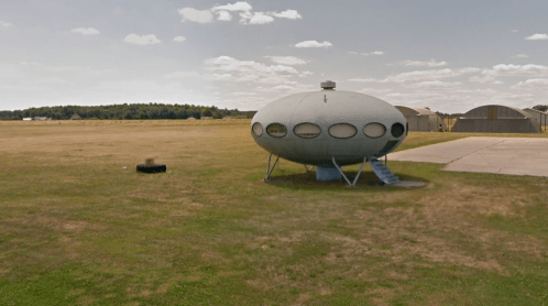 A futuristic, oval-shaped building on a grassy field, with a staircase and nearby hangars in the background.