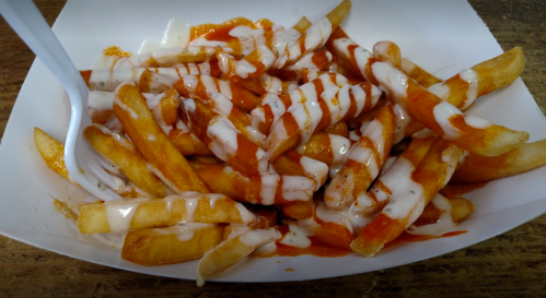 A serving of French fries drizzled with hot sauce and creamy dressing in a white container.