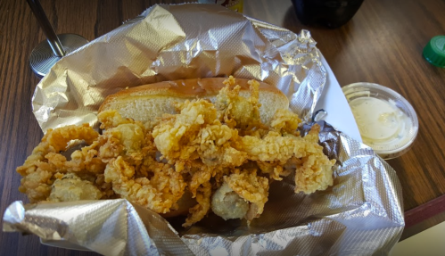 A fried seafood sandwich with crispy fried oysters on a bun, served in foil with a side of dipping sauce.