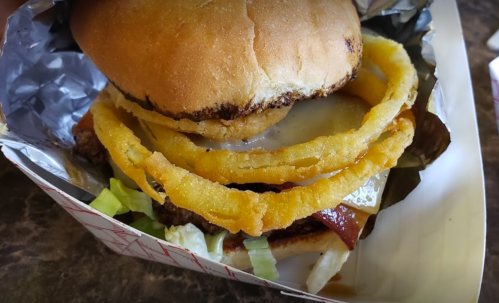 A close-up of a burger with melted cheese, lettuce, and crispy onion rings in a toasted bun, wrapped in foil.