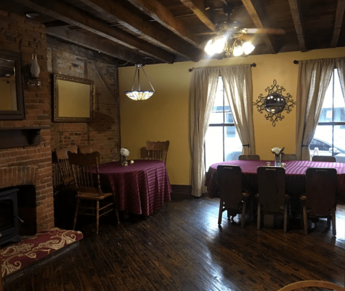 Cozy dining room with wooden beams, brick walls, and tables set with maroon tablecloths and floral centerpieces.