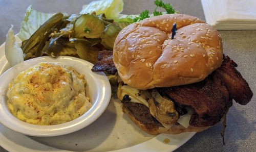 A sesame seed bun burger with bacon, cheese, and mushrooms, served with potato salad and pickles on the side.