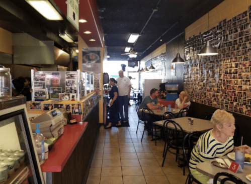 A busy diner interior with a counter, customers seated, and a wall covered in photos. Staff are interacting with patrons.