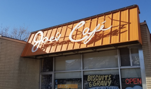 Sign for Joe's Café with an orange awning, featuring "Biscuits & Gravy" and an "Open" neon sign in the window.