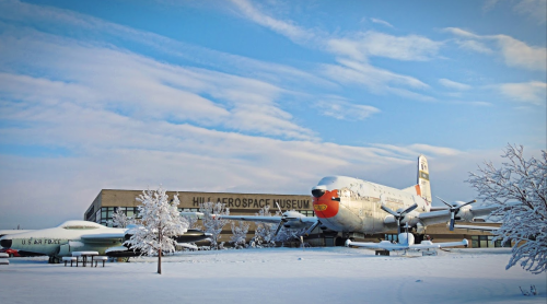A snowy scene featuring the Hill Aerospace Museum with a vintage airplane displayed outside.