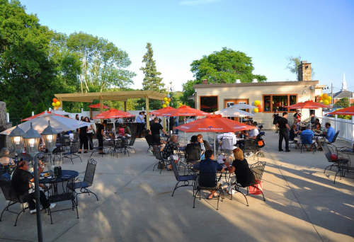 Outdoor gathering at a venue with tables, umbrellas, and guests socializing in a sunny setting.