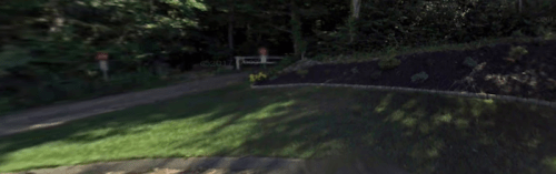 A wooded area with a path, grassy lawn, and a flower bed, partially shaded by trees.